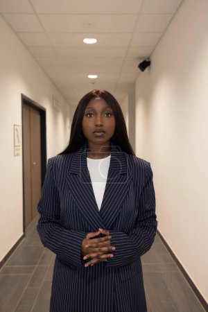 Ce portrait captivant montre une jeune femme noire debout dans un couloir de bureau, qui respire la confiance et l'autorité. Elle est vêtue d'un blazer à rayures marine sur un haut blanc croustillant.
