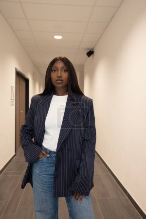 This portrait depicts a young Black woman in a professional setting, exhibiting a blend of sophistication and casual style. She stands confidently in a corridor, wearing a navy blue pinstriped blazer