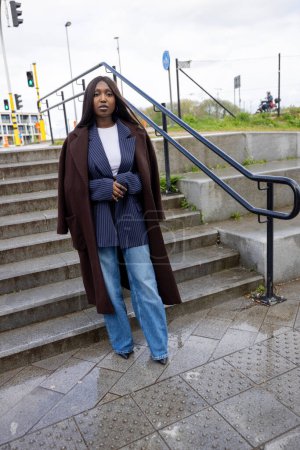 Este retrato dinámico captura a una joven mujer negra de pie con confianza en las escaleras de la ciudad, vestida con un elegante atuendo en capas. Lleva un largo abrigo marrón sobre una chaqueta azul marino de rayas blancas emparejada con