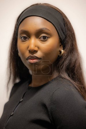 This close-up portrait captures a young Black woman with a serene expression, dressed in a classic black top accessorized with a simple headband and elegant earrings. Her gaze is direct and engaging