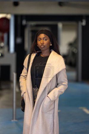 This portrait captures a young Black woman exuding sophistication and grace, dressed in a stylish light beige coat over a chic black outfit. Standing in front of an urban business entrance, her poised
