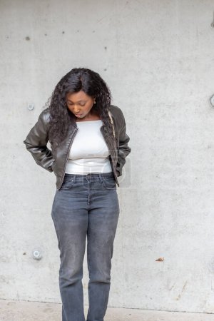 This portrait features a contemplative African American woman standing against a textured concrete wall. Dressed in a stylish leather jacket and casual jeans, her pose and downward gaze suggest