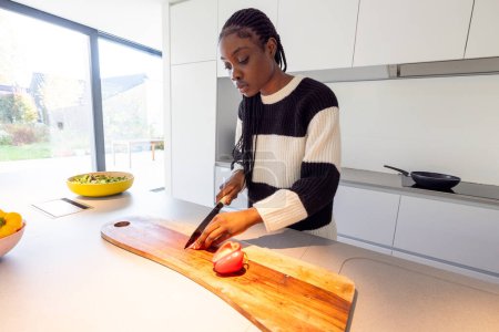 This image captures a young Black woman focused on slicing a tomato on a wooden cutting board in a well-lit, modern kitchen. She is dressed in a casual black and white striped sweater. The kitchen is