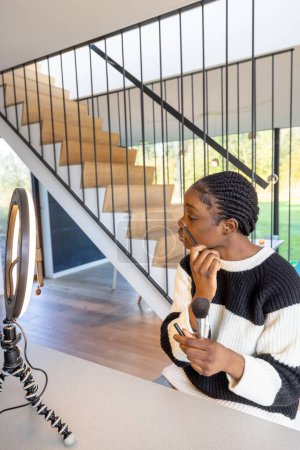 This image features a young Black woman engaging in her morning beauty routine, applying makeup while seated at a modern vanity table. The setting is stylish, with a well-lit interior and a sleek