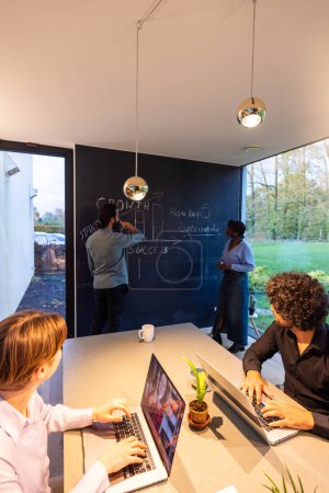 This image captures a diverse team of professionals engaged in a brainstorming session in a contemporary office setting. Two individuals, a Black woman and a man, are discussing strategies written on