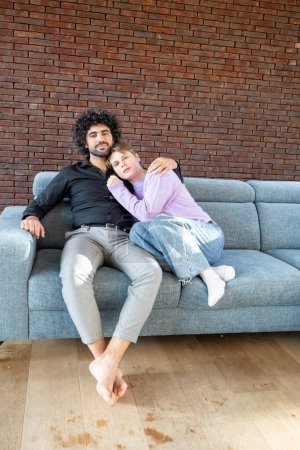 This heartwarming image showcases a multicultural couple enjoying a relaxed moment together on a grey sofa, set against a stylish brick wall background. The man, with curly hair and casual attire
