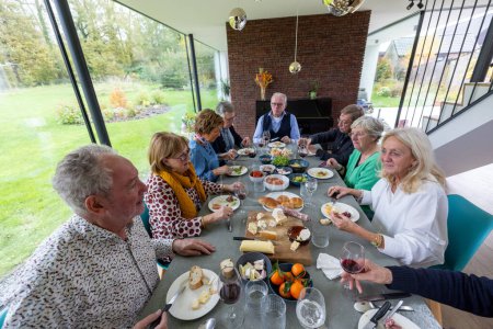 Une scène vibrante d'un groupe d'amis âgés réunis autour d'une table à manger, profitant d'un repas festif dans une maison moderne. Le groupe, composé d'hommes et de femmes, est engagé dans une conversation animée