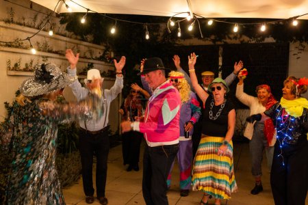 A diverse group of elderly individuals is seen having a joyous time at an outdoor costume party. The attendees, both men and women, are dressed in colorful and extravagant costumes, including cowboy