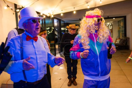 Eine einnehmende Szene fängt ältere Männer in Kostümen ein, die eine lebhafte Outdoor-Party genießen. Ein Mann trägt einen weißen Cowboyhut, eine Sonnenbrille und eine rote Fliege, während ein anderer einen lebhaften Retro-Look trägt.