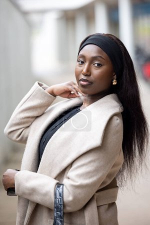 This portrait captures a young Black woman striking a confident pose on a city street, dressed in a stylish beige coat over a black ensemble. Her relaxed posture, combined with a thoughtful expression
