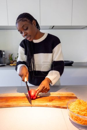 This image captures a young Black woman focused on preparing a healthy meal in a modern kitchen. She is slicing a tomato with care on a wooden chopping board, next to a bowl of freshly grated carrots