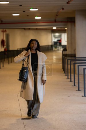 This image captures a young Black woman walking confidently through an urban indoor setting, dressed in a chic winter ensemble. She wears a light beige coat over a black outfit, accessorized with a