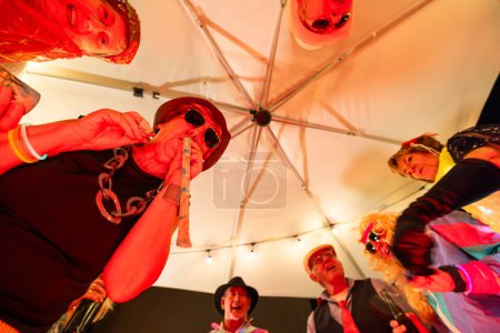 A dynamic close-up shot captures a group of elderly friends enthusiastically blowing party horns at an outdoor costume party. The image is taken from a low angle, emphasizing the large white canopy