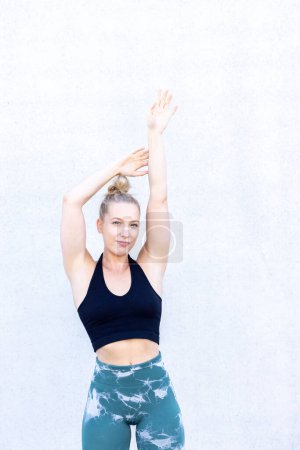 A woman in athletic wear stands with one arm extended upwards, suggesting a yoga pose. The background is plain and untextured, focusing attention on her balanced pose and fit physique. Active Woman