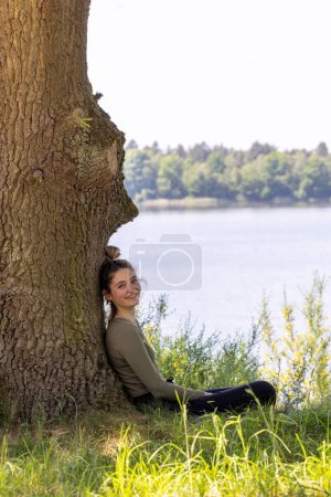 Captured in a serene natural setting, a young woman leans against a sturdy tree, her smile reflecting a moment of relaxation. The lake in the background and the greenery around her contribute to the