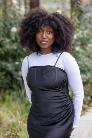 This image features a Black woman standing outdoors, wearing a stylish black dress over a long-sleeve white top. Her natural, curly hair frames her face, and she gazes confidently at the camera with a