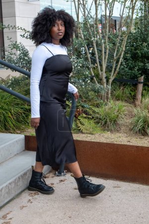 This image features a Black woman descending outdoor stairs in an urban setting, dressed in a stylish black dress layered over a long-sleeve white top, paired with black combat boots. Her natural