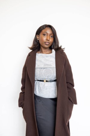 This image features a Black woman posing against a white background, dressed in a stylish brown coat over a gray sweater and a black skirt. Her hair is straight and shoulder-length, framing her face