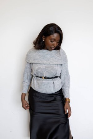 This image features a Black woman posing against a white background, dressed in a stylish gray sweater and a black skirt. She is looking downwards with a thoughtful expression, adding a contemplative