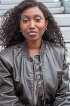 This image features a Black woman posing outdoors, wearing a stylish leather jacket. Her curly hair cascades around her face, and she gazes confidently at the camera. She accessorizes with gold hoop