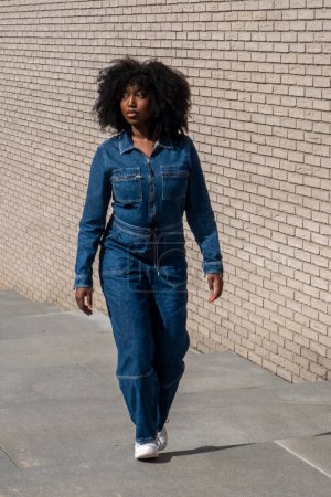 Dieses Bild zeigt eine schwarze Frau, die selbstbewusst im Freien an einer Ziegelmauer entlang geht. Sie trägt einen stylischen Jeansanzug gepaart mit weißen Turnschuhen, und ihr natürliches, lockiges Haar umrahmt ihr Gesicht.