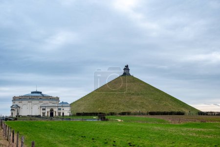 Cette photographie représente le monticule des Lions avec son imposante statue de lion surplombant le terrain, à côté du centre d'accueil du champ de bataille historique de Waterloo. Les champs verts tentaculaires dans le