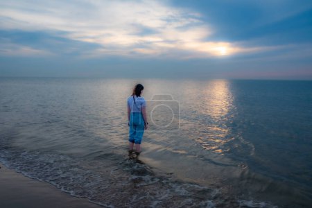 Das Bild fängt eine einsame Gestalt ein, die in der ruhigen See steht und in Richtung der Sonne schaut, während sie in der weiten Horizontlinie untergeht. Die sanften Wellen des Meeres umschlingen ihre hochgekrempelten Jeans, was auf eine
