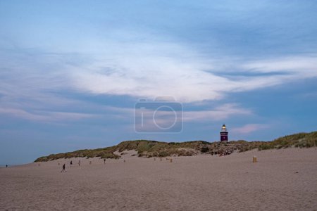 Im weichen Licht des frühen Abends spazieren mehrere Personen gemütlich am ausgedehnten Sandstrand entlang, im Hintergrund erheben sich die Dünen sanft. Ein markanter Leuchtturm, dessen lebhafte