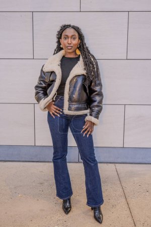 Stylish woman confidently posing in leather jacket and jeans by modern building wall, showcasing urban fashion