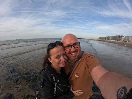 A couple happily poses for a selfie on a sandy beach with an urban skyline in the background on a sunny day