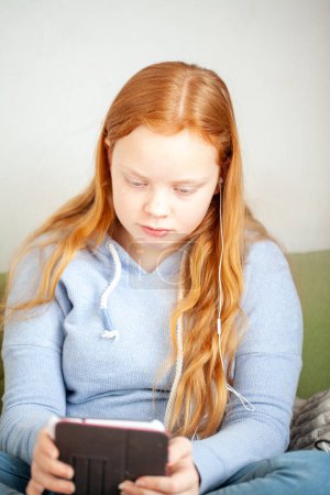 Una adolescente con el pelo rojo está sentada en un sofá, usando una tableta y auriculares, apareciendo enfocada y concentrada