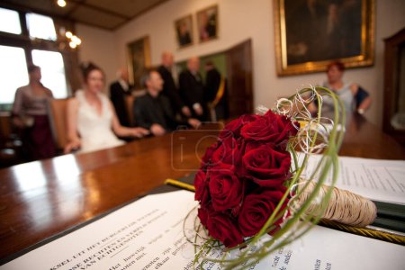 Red rose wedding bouquet on table in registry office, wedding ceremony attendees blurred in the background.