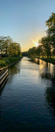A serene canal with calm waters reflects the sunset light, surrounded by trees, creating a peaceful evening scenery