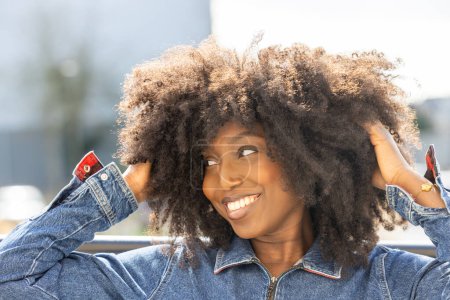 A cheerful woman with curly afro hair in a denim jacket is smiling happily outdoors under the sunshine