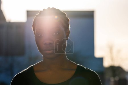 Una mujer afroamericana serena está de pie en una silueta, retroiluminada por una llamativa llamarada solar que crea un efecto halo alrededor de su cabeza. Su expresión equilibrada y tranquila es parcialmente visible, ofreciendo una