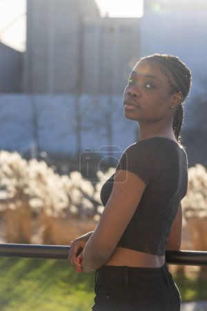 Photo for This image presents a young African woman turned slightly over her shoulder, giving a confident look towards the camera. The background features an out-of-focus urban landscape with industrial - Royalty Free Image