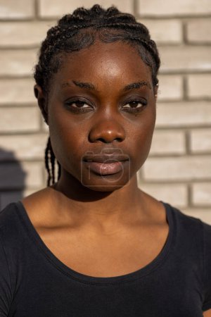 A young African womans portrait is set against a white brick wall, with the shadows of the surrounding environment creating a pattern across her face. The sunlight highlights her hair braids, the