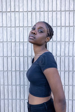 Ein afrikanisches Frauenporträt vor dem Hintergrund weißer geometrischer Kacheln, das einen Moment der Reflexion nach oben einfängt. Das schwarze Crop Top und die Jeans, die sie trägt, ergänzen ihre Figur, während ihre Zöpfe