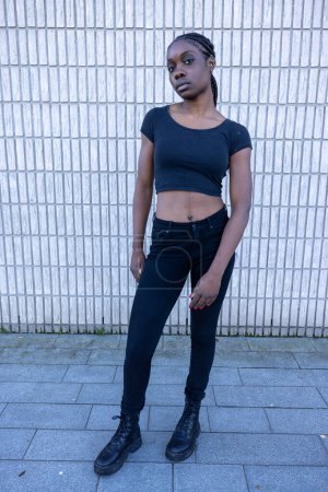 Dieses Foto zeigt eine junge Afrikanerin, die selbstbewusst vor einer weißen Backsteinwand steht. Bekleidet ist sie mit einem formschlüssigen schwarzen bauchfreien Oberteil und Jeans mit hohem Bund, dazu klobige Schnürstiefel.