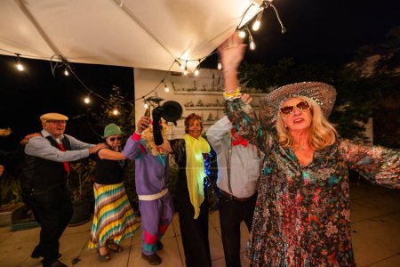 A lively group of elderly friends is captured dancing and celebrating at an outdoor costume party under festive string lights and a large white canopy. The attendees, both men and women, are dressed