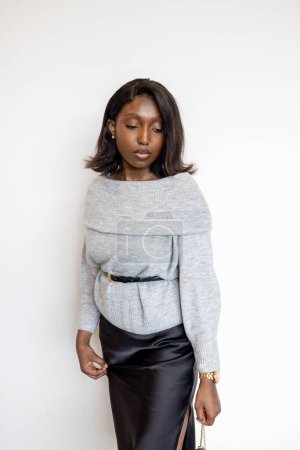 This portrait features a stylish young Black woman standing against a plain white background. She is dressed in an off-the-shoulder grey sweater, cinched at the waist with a black belt, and a sleek