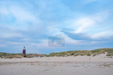 This image presents a serene beachscape, featuring undulating coastal dunes topped with hardy dune grasses. Standing prominently in the background is a striking lighthouse, its deep hue contrasting