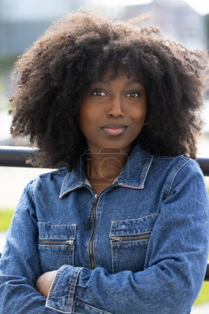 A confident woman with natural curls poses outdoors in a denim jacket, exuding a stylish and modern vibe