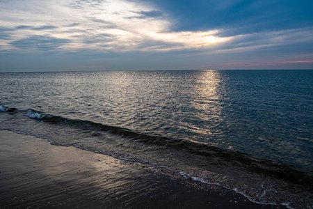 Esta imagen es una serena representación de la puesta de sol en la playa, con el reflejo de los soles brillando a través de las suaves olas del océano. El cielo, una mezcla de grises suaves y azules con un toque de naranja cálido de la