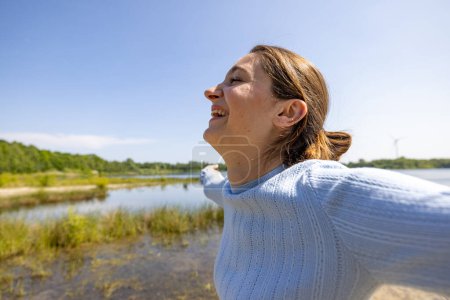 Une femme en pull bleu regarde joyeusement le paysage calme au bord du lac, immergée dans la tranquillité de la nature