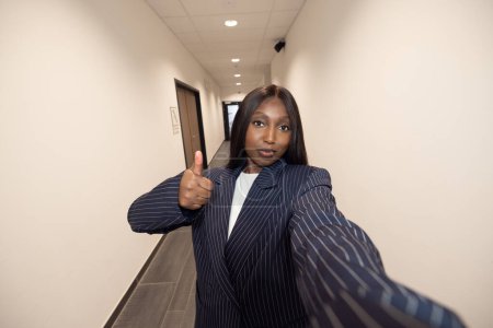 Femme d'affaires confiante dans un costume à rayures prendre un selfie dans un couloir, donnant un pouce geste
