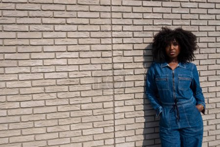 Fashionable woman with curly hair, in denim, stands outdoors against a brick wall exuding style and confidence