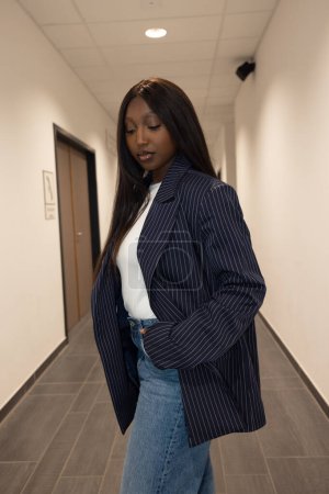 A stylish, confident woman in a pinstripe blazer and denim jeans striking a pose in a contemporary hallway