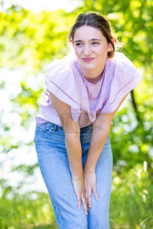 A young woman is pictured bending forward, hands on knees, in a playful stance amidst a lush green backdrop. Her expression is one of amused delight, with a hint of a mischievous smile. The light
