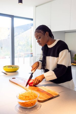 Une jeune femme tranche des légumes sur une planche à découper en bois dans une cuisine moderne inondée de lumière naturelle